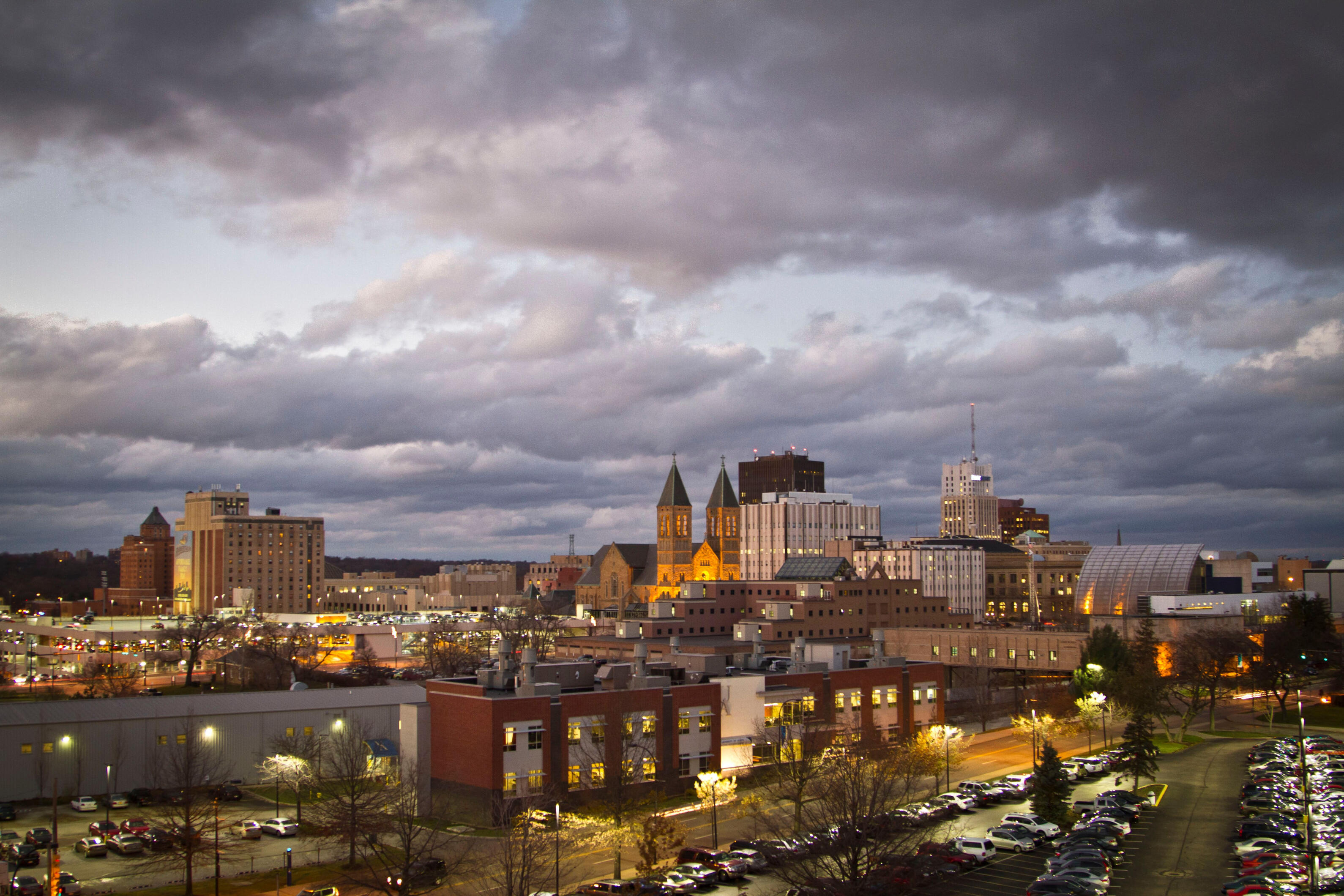 Downtown Akron, Ohio skyline at dusk, with historic church towers, illuminated buildings, and dramatic clouds overhead.